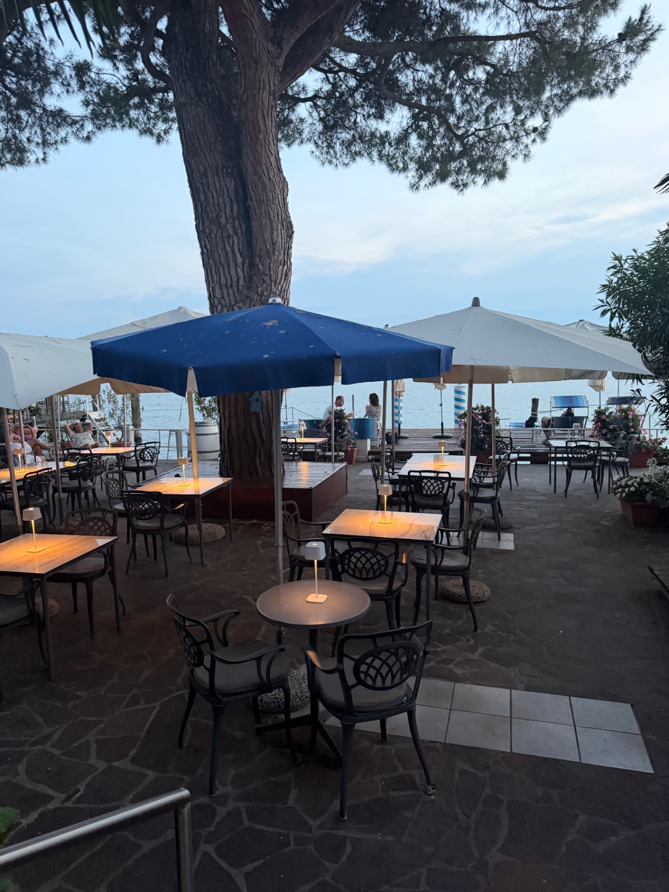 Lakeside hotel terrace at dusk with warm table lamps, parasols and a pine tree overlooking Lake Garda